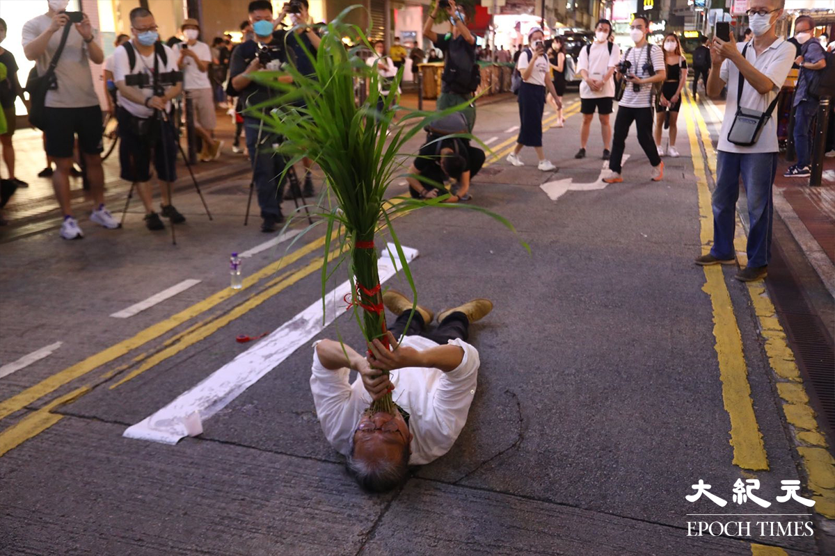 穿著白恤衫的男行為藝術家，以紅絲帶捆綁雜草底端，狀似在拉小提琴。之後，他躺在地上將雜草放進口中，把身體成十字架形躺著。（胡琪／大紀元）